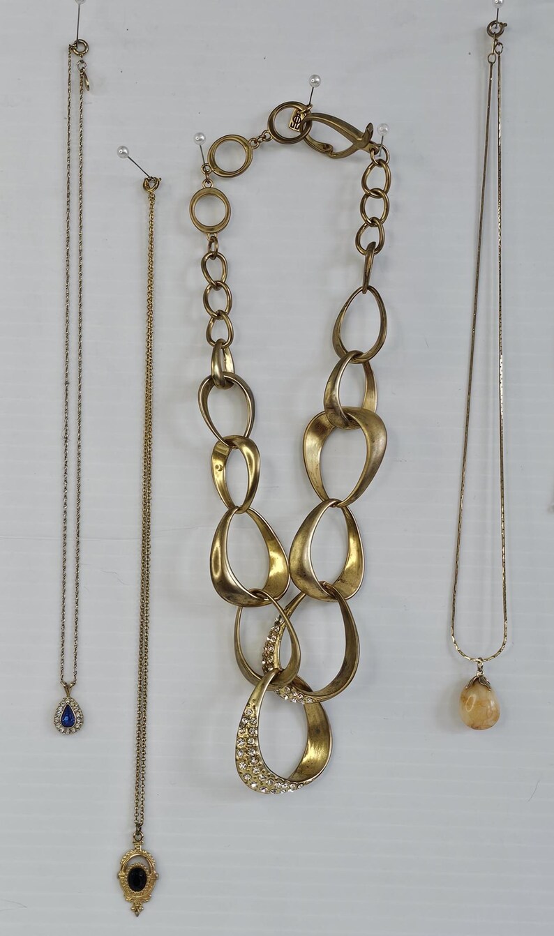 May include: A collection of gold-toned necklaces, each with unique designs. One features a large, linked chain with a crystal-studded pendant. Others include delicate chains with teardrop and oval pendants. The necklaces are displayed against a white background.