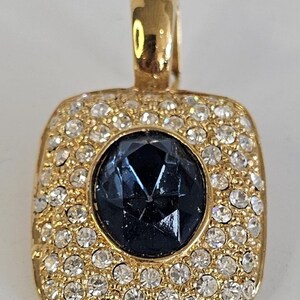 May include: A gold-toned pendant with a square shape, encrusted with small, clear crystals around a central, faceted, dark blue gemstone. The pendant has a loop at the top for a chain.