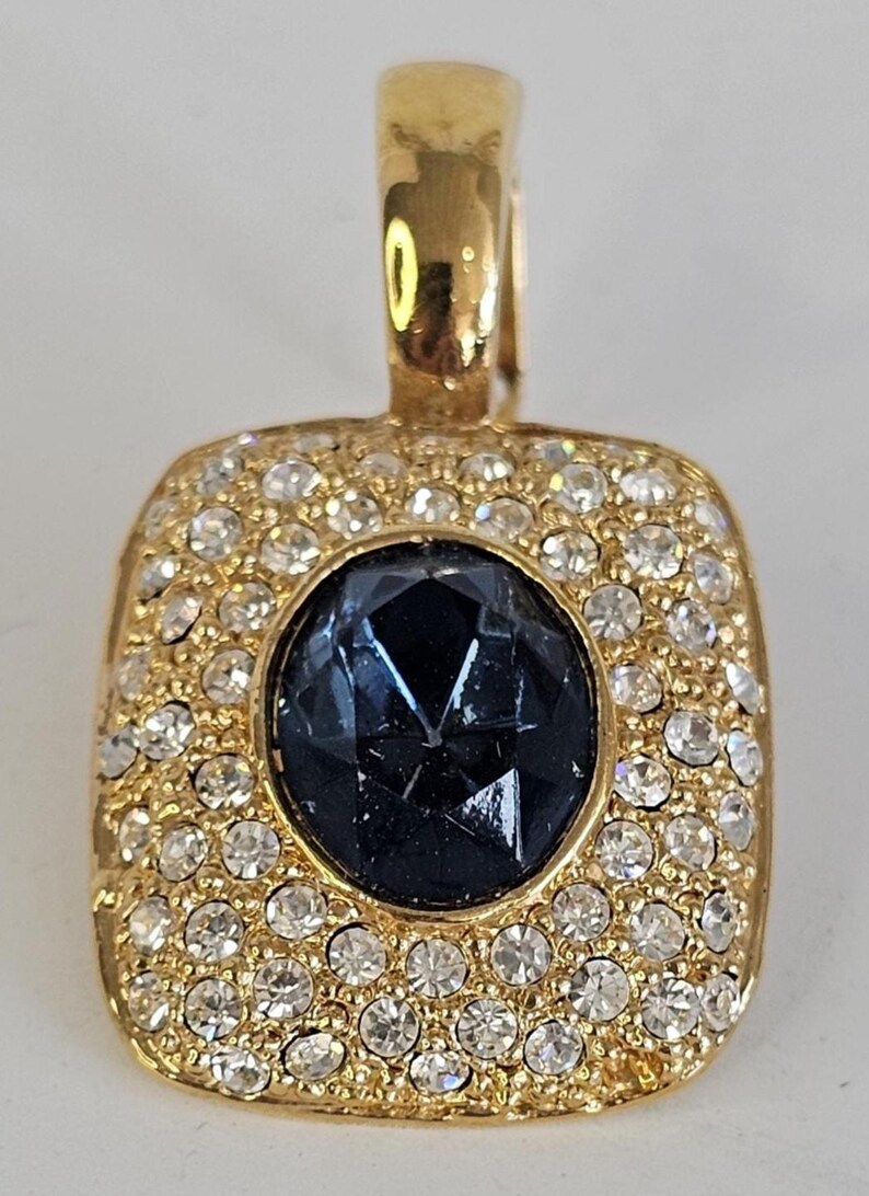 May include: A gold-toned pendant with a square shape, encrusted with small, clear crystals around a central, faceted, dark blue gemstone. The pendant has a loop at the top for a chain.