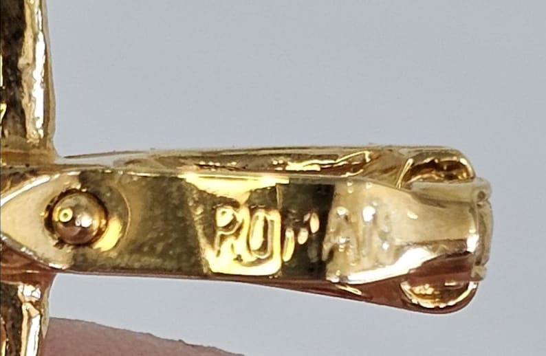 May include: Close-up of a gold-colored jewelry clasp with the engraved text "ROMA". The clasp has a detailed design and a shiny finish, suggesting a vintage or high-end piece. The background is a neutral color.