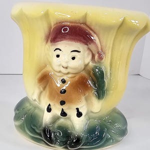 May include: A vintage ceramic planter in the shape of a yellow flower with a smiling gnome figure. The gnome wears a red hat and brown coat with black buttons. The planter sits on a green base.