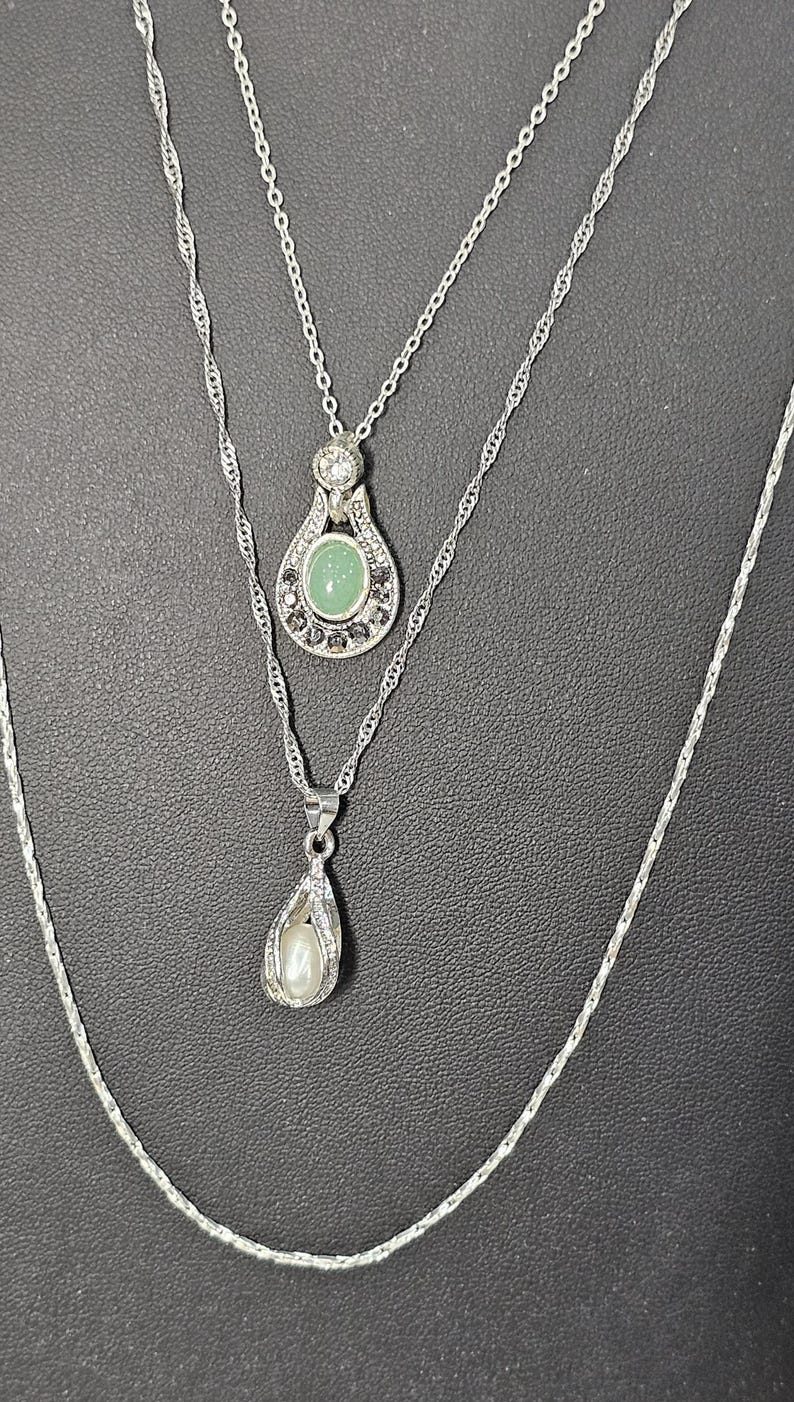 Peut inclure: Un collier argent&eacute; &agrave; trois rangs avec trois pendentifs. Le pendentif sup&eacute;rieur pr&eacute;sente une pierre verte entour&eacute;e de pierres plus petites. Le pendentif du milieu est en forme de larme avec une perle &agrave; l'int&eacute;rieur. Le pendentif inf&eacute;rieur est une simple cha&icirc;ne en argent.