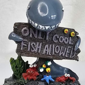 May include: A whimsical shark-themed figurine with a sign that reads "ONLY COOL FISH ALLOWED." The shark is gray with a wide grin, and the sign is made of wood-like material. The base features red crabs, a blue starfish, and green plants.