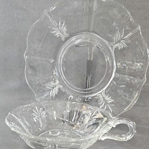 May include: Clear glass teacup and saucer set. The teacup has a handle and a scalloped rim. The saucer is round with a scalloped edge and a raised center. Both pieces feature a leaf design.
