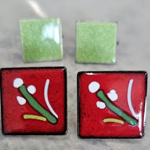 May include: Two pairs of square earrings. The top pair features a solid light green textured finish. The bottom pair is red with a graphic design consisting of a green stem, white circular accents, and a small yellow horizontal line.