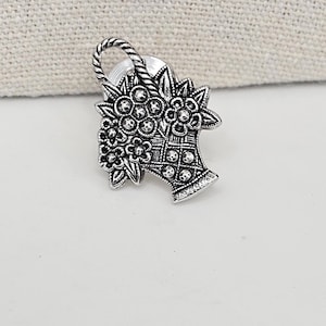 May include: A silver-toned metal brooch in the shape of a basket overflowing with flowers. The basket and flowers are intricately detailed with a textured finish.