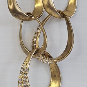 May include: A gold-toned necklace featuring a series of interlocking teardrop-shaped links. Some links are embellished with small, sparkling crystals, adding a touch of glamour to the jewelry. The necklace has a vintage or antique aesthetic.