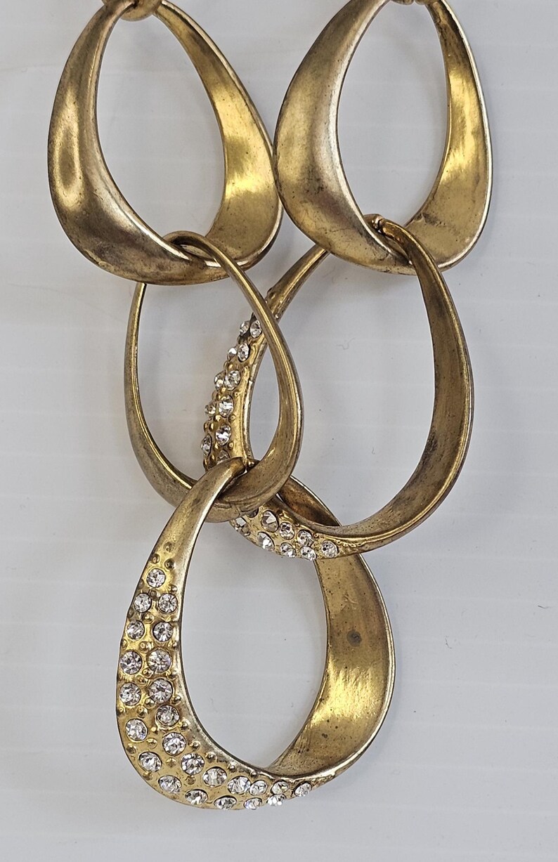 May include: A gold-toned necklace featuring a series of interlocking teardrop-shaped links. Some links are embellished with small, sparkling crystals, adding a touch of glamour to the jewelry. The necklace has a vintage or antique aesthetic.