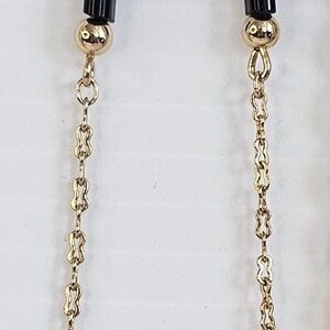 May include: Gold-toned necklace featuring a repeating pattern of small, figure-eight links. The necklace has black, rectangular beads and gold spherical beads. The necklace is displayed against a white background.
