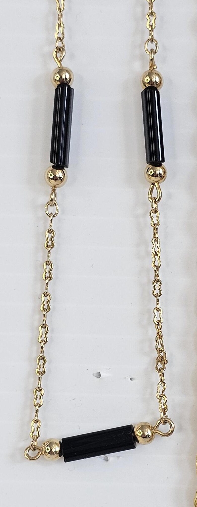 May include: Gold-toned necklace featuring a repeating pattern of small, figure-eight links. The necklace has black, rectangular beads and gold spherical beads. The necklace is displayed against a white background.