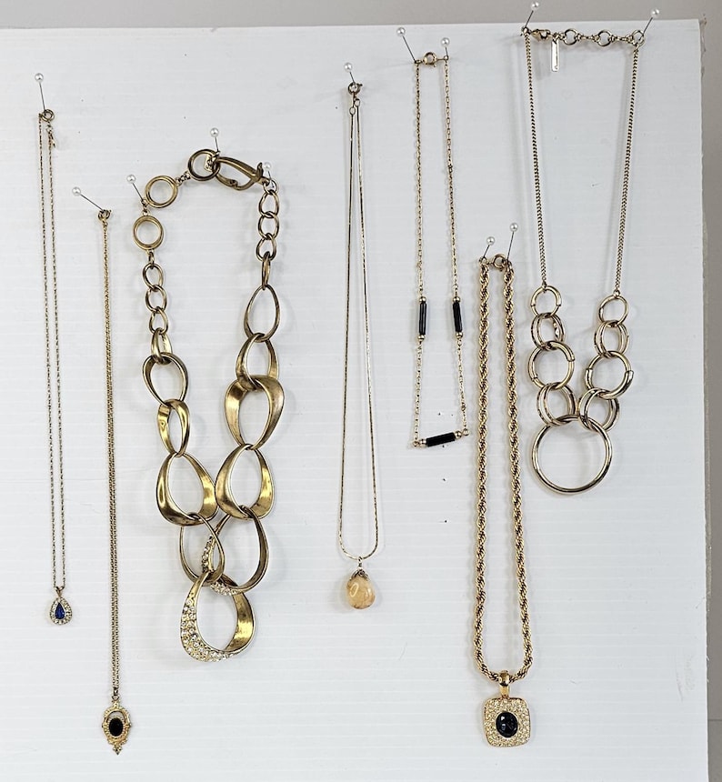 May include: A collection of gold-toned necklaces displayed against a white background. The necklaces vary in style, including delicate chains with pendants, chunky link designs, and rope chains. Some feature gemstones or black accents.