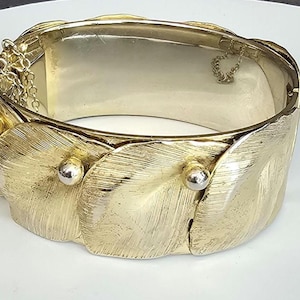 May include: A gold-toned hinged bangle bracelet with a textured leaf design. The bracelet features a rectangular interior and is secured with a clasp and safety chain. Silver-colored accents are visible on the bracelet.