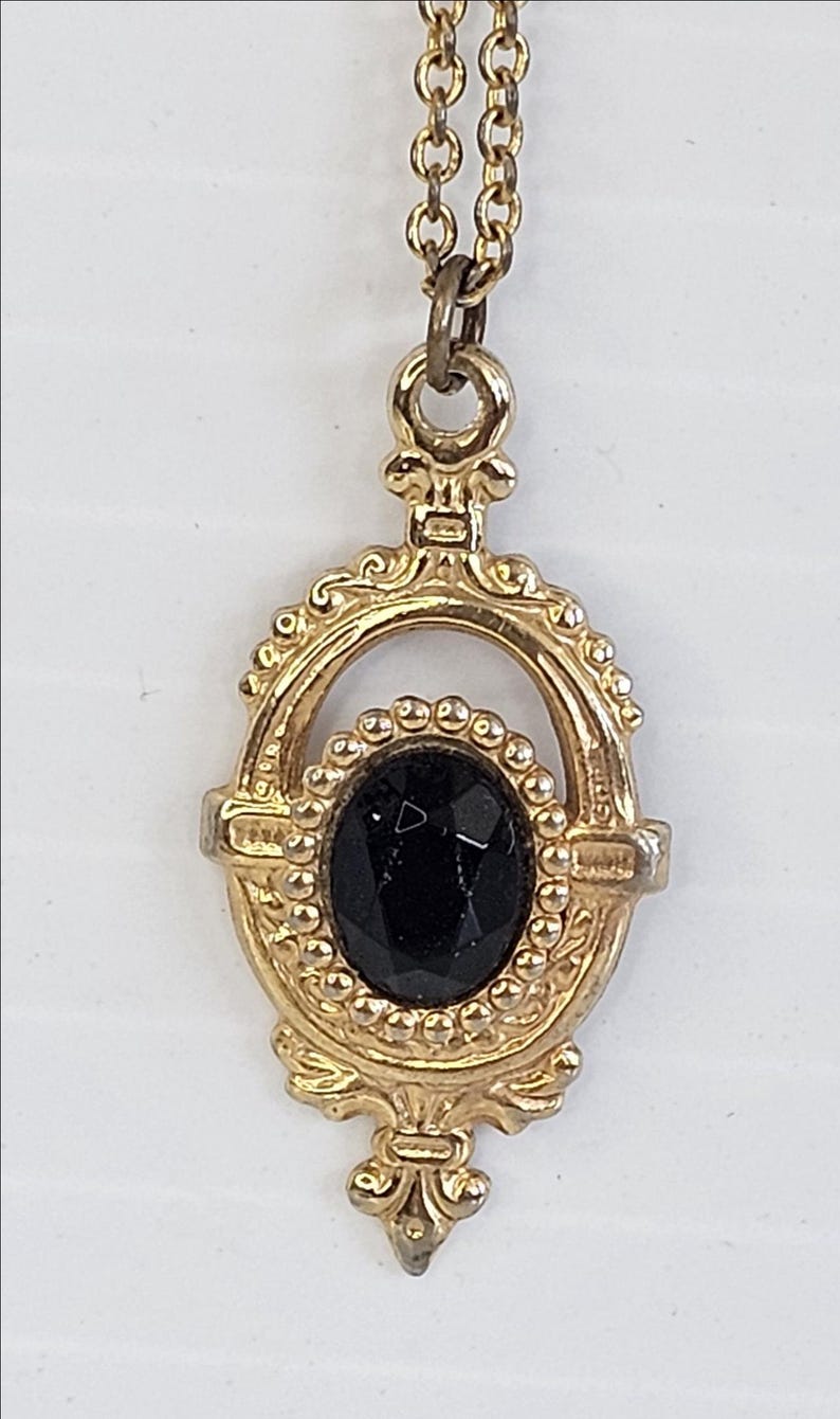 May include: A gold-toned pendant necklace featuring an oval black gemstone. The pendant has an ornate, vintage design with a decorative frame and a delicate chain.