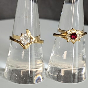 May include: Two gold rings displayed on clear stands. One ring features a large, clear gemstone with smaller accent stones and a floral design. The other ring has a red center stone surrounded by smaller clear stones in a flower shape.