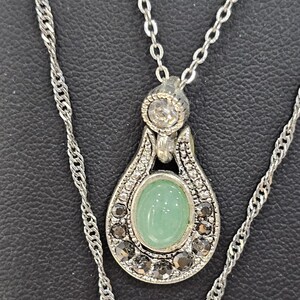 Peut inclure: Un collier en cha&icirc;ne argent avec un pendentif orn&eacute; d'une pierre ovale verte entour&eacute;e de petites pierres claires et noires.