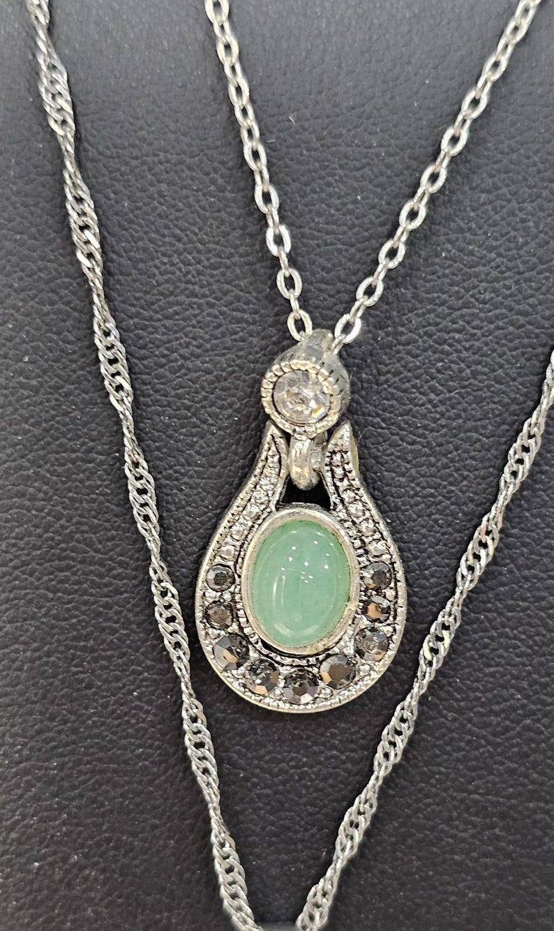 Peut inclure: Un collier en cha&icirc;ne argent avec un pendentif orn&eacute; d'une pierre ovale verte entour&eacute;e de petites pierres claires et noires.
