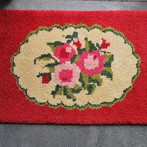 May include: A rectangular rug with a vibrant red background and a central cream-colored oval medallion. The medallion features a detailed floral pattern with pink and red roses, green leaves, and small red buds, all rendered in a textured loop pile.