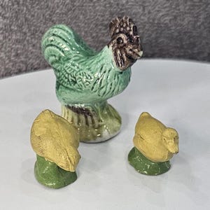 May include: A green and brown ceramic rooster figurine with two smaller yellow ceramic chicks. The rooster has a detailed design and the chicks have simple designs.