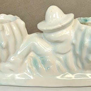 May include: A vintage ceramic planter in a light blue and white color scheme. The planter features two open containers on either side of a reclining figure wearing a hat. The planter is a decorative item.