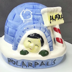 May include: A ceramic figurine of an igloo with a smiling face peeking out. The igloo is blue with white accents, and the base reads "POLAR PALS." A signpost with "ALASKA" is next to the igloo, with red and white stripes.