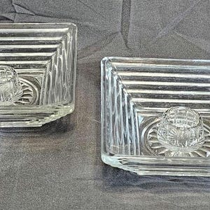 May include: Two clear glass candle holders. Each square holder has a central circular candle cup and a geometric design. The holders are on a gray surface.