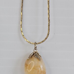 May include: A gold-colored chain necklace with a teardrop-shaped pendant. The pendant is a light beige color with a marbled appearance, and is held by a silver-colored metal clasp.