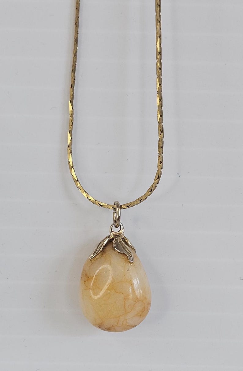 May include: A gold-colored chain necklace with a teardrop-shaped pendant. The pendant is a light beige color with a marbled appearance, and is held by a silver-colored metal clasp.