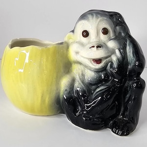 May include: A ceramic planter in the shape of a smiling gorilla. The gorilla is black and white with brown eyes and a red mouth. The planter section is yellow and egg-shaped.