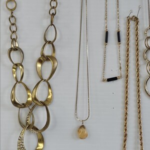 May include: A collection of gold-toned necklaces, including a chunky chain link necklace, a delicate chain with a teardrop pendant, and a rope chain with a square pendant. Some necklaces have gemstone accents.