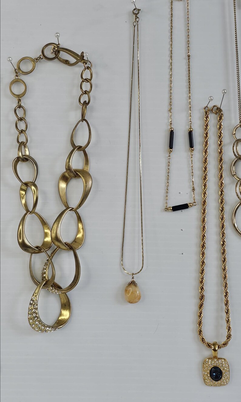 May include: A collection of gold-toned necklaces, including a chunky chain link necklace, a delicate chain with a teardrop pendant, and a rope chain with a square pendant. Some necklaces have gemstone accents.