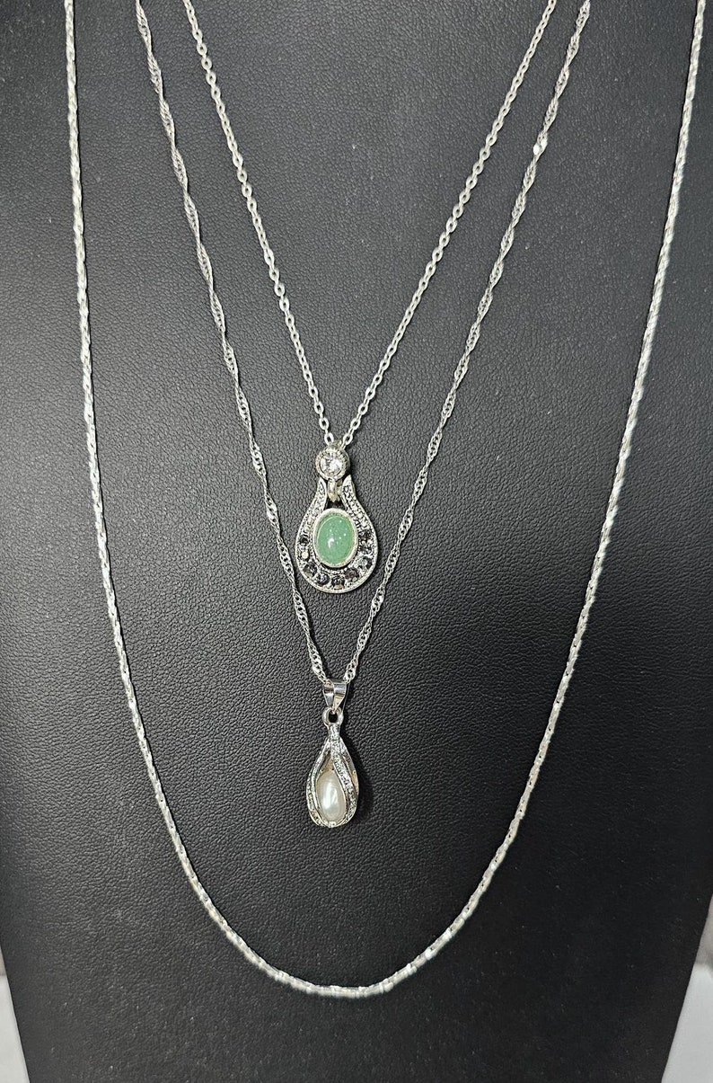 Peut inclure: Un collier argent&eacute; &agrave; plusieurs rangs avec deux pendentifs. Le pendentif sup&eacute;rieur pr&eacute;sente une pierre pr&eacute;cieuse verte entour&eacute;e de pierres plus petites. Le pendentif inf&eacute;rieur pr&eacute;sente une perle blanche.