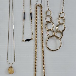 May include: A collection of gold-toned necklaces, each with a unique design. One features a teardrop pendant, another has black bar accents, and a third is a rope chain with a square pendant. The necklaces are displayed against a white background.