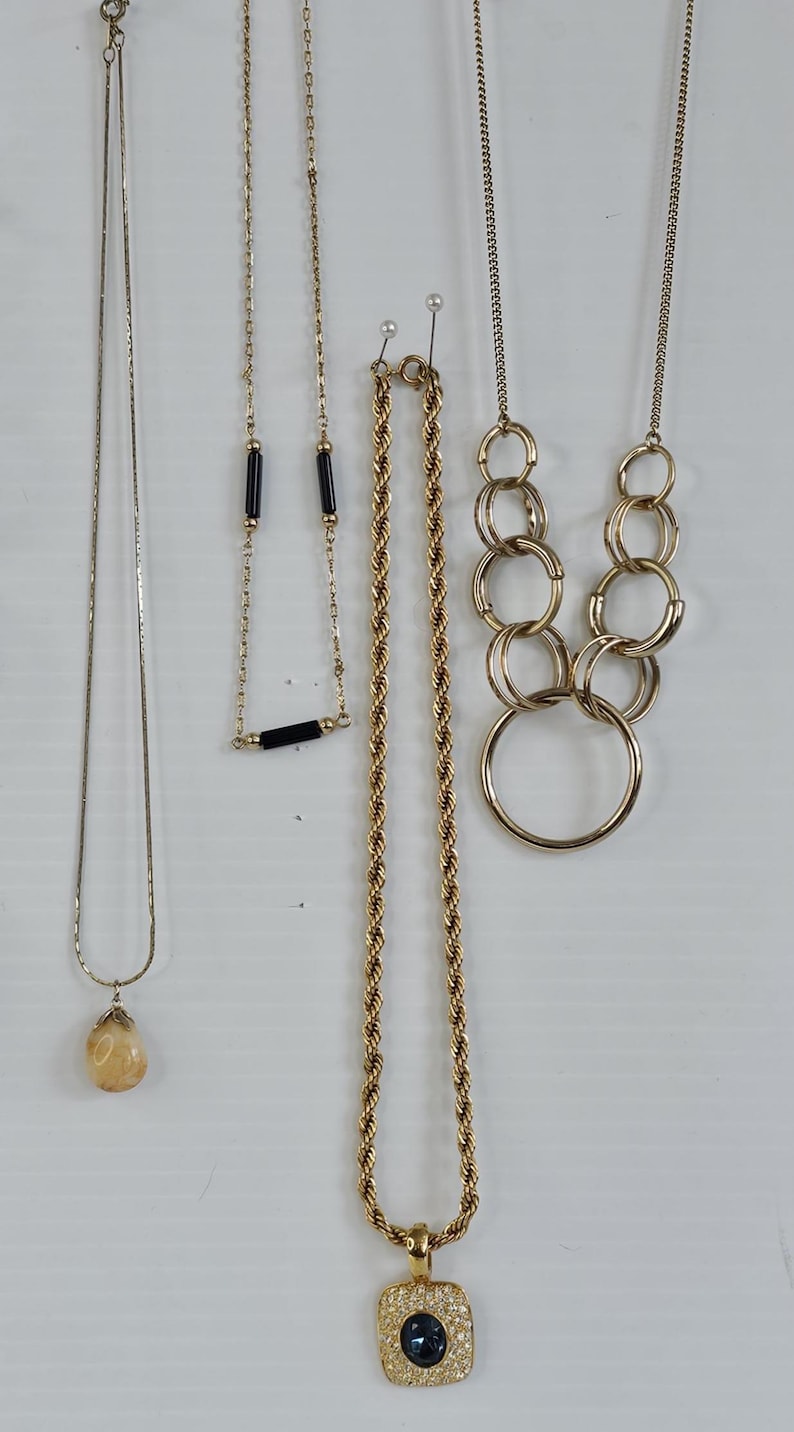 May include: A collection of gold-toned necklaces, each with a unique design. One features a teardrop pendant, another has black bar accents, and a third is a rope chain with a square pendant. The necklaces are displayed against a white background.