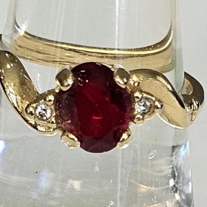 May include: A gold ring featuring a large, oval-cut, deep red gemstone as the centerpiece. Two smaller clear gemstones flank the main stone. The ring has a swirling design on the band.