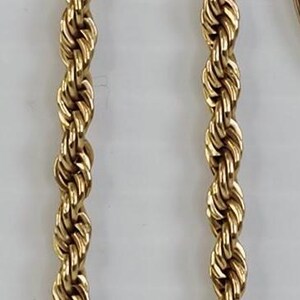 May include: A gold-toned necklace with a rope chain design. The necklace features a rectangular pendant with a dark blue center stone surrounded by clear crystals. The necklace is displayed on a white background.