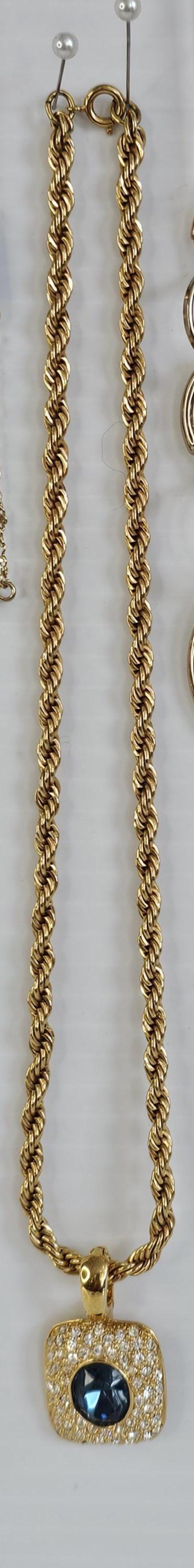 May include: A gold-toned necklace with a rope chain design. The necklace features a rectangular pendant with a dark blue center stone surrounded by clear crystals. The necklace is displayed on a white background.