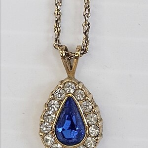 May include: Gold-toned pendant necklace featuring a teardrop-shaped blue gemstone surrounded by small, clear crystals. The pendant hangs from a delicate gold chain, creating an elegant and classic jewelry piece.