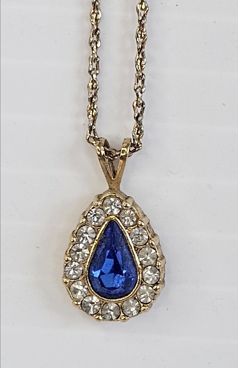 May include: Gold-toned pendant necklace featuring a teardrop-shaped blue gemstone surrounded by small, clear crystals. The pendant hangs from a delicate gold chain, creating an elegant and classic jewelry piece.