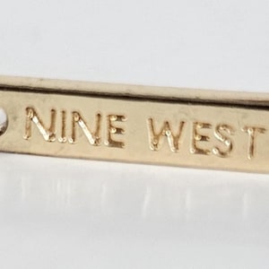 May include: Close-up of a gold-colored jewelry tag with the engraved text "NINE WEST". The tag is rectangular with rounded edges and is attached to a chain link. The background is a light, neutral color.