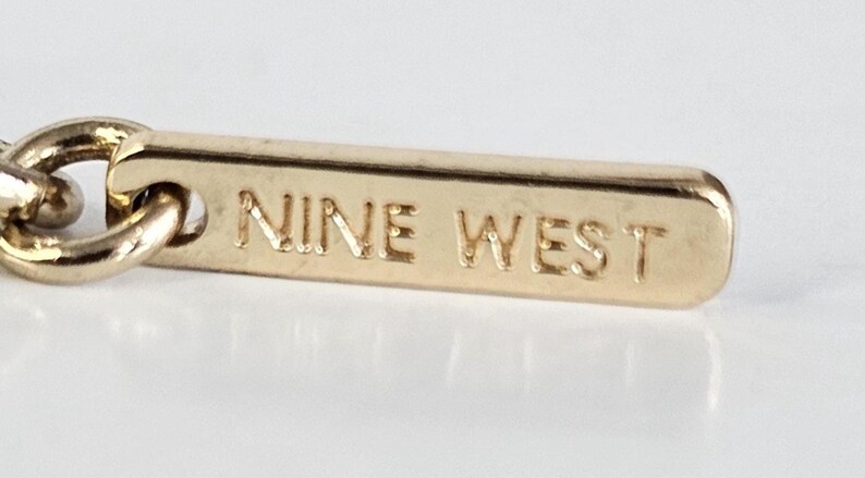 May include: Close-up of a gold-colored jewelry tag with the engraved text "NINE WEST". The tag is rectangular with rounded edges and is attached to a chain link. The background is a light, neutral color.