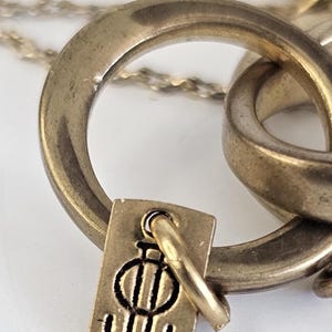 May include: Close-up of a gold-toned necklace featuring a pendant with a stylized design. The pendant is rectangular with a small loop attached to a larger ring. The necklace chain is visible in the background.