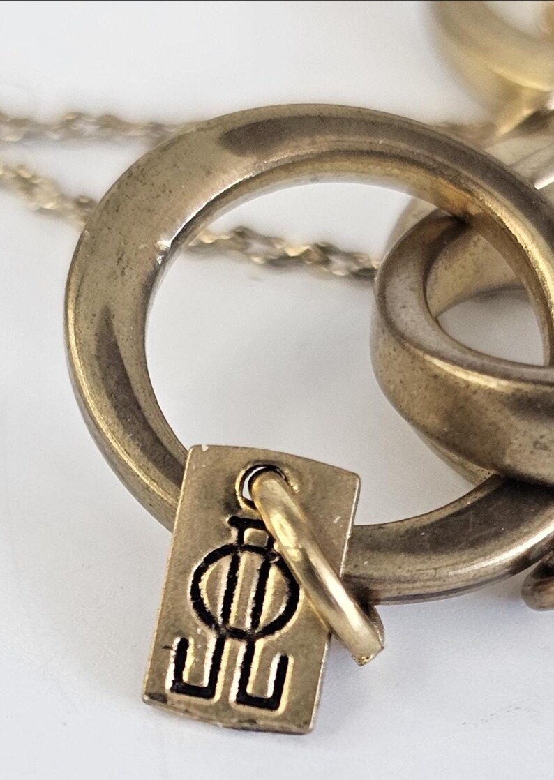 May include: Close-up of a gold-toned necklace featuring a pendant with a stylized design. The pendant is rectangular with a small loop attached to a larger ring. The necklace chain is visible in the background.