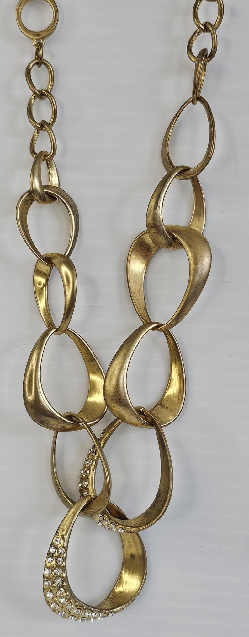 May include: Gold-toned statement necklace featuring large, interconnected oval links. One link is embellished with sparkling rhinestones. The necklace has a vintage aesthetic and is suitable for special occasions or adding a touch of glamour to any outfit.