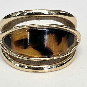 May include: Gold-tone ring with a tortoise shell-style oval center. The ring features a double-band design, with the tortoise shell element set between the bands. The ring is a classic jewelry piece.