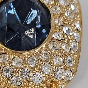 May include: Close-up of a gold-toned, square-shaped brooch. The brooch features a large, faceted, dark blue gemstone at its center, surrounded by a border of small, clear crystals. The crystals are set in a gold-colored metal.