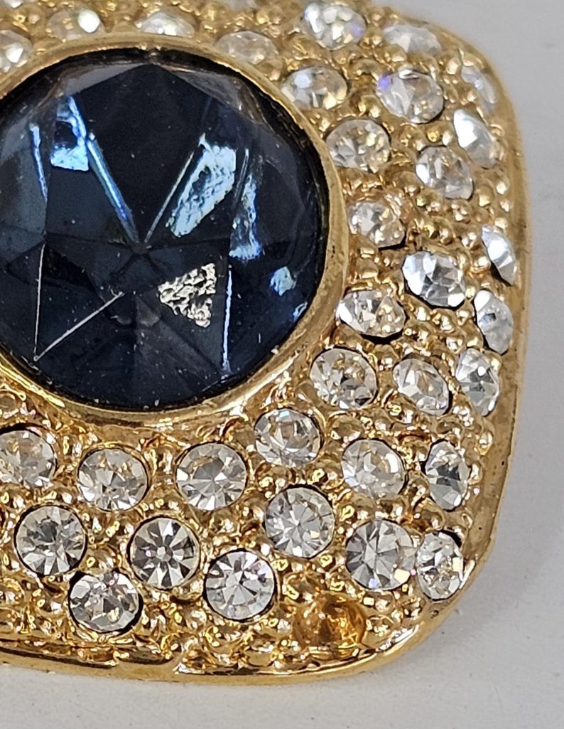 May include: Close-up of a gold-toned, square-shaped brooch. The brooch features a large, faceted, dark blue gemstone at its center, surrounded by a border of small, clear crystals. The crystals are set in a gold-colored metal.