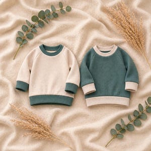 May include: Two baby sweatshirts. One is cream with teal trim, the other is teal with cream trim. Both have long sleeves and a crew neck. The sweatshirts are displayed on a beige, textured surface with decorative sprigs.