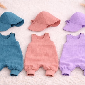 Puede incluir: Tres conjuntos de bebé en azul, melocotón y lavanda, cada uno con un sombrero para el sol a juego. Los conjuntos son peleles de tela texturizada. Decoraciones de madera en forma de estrella y nube están dispersas.