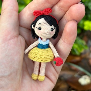 May include: A crocheted doll of Snow White, wearing a yellow dress with a blue and white bodice, a red bow, and holding a red apple.