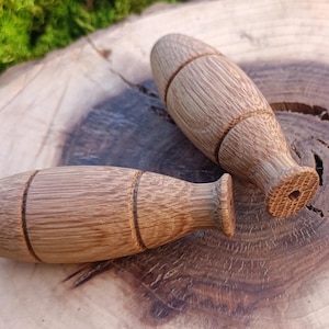 Può includere: Due tappi per bottiglia in legno con finitura marrone chiaro e linee di accento marrone scuro. I tappi hanno una forma arrotondata a forma di botte con una base svasata e un foro al centro. Sono esposti su una fetta di legno con uno sfondo di muschio verde.